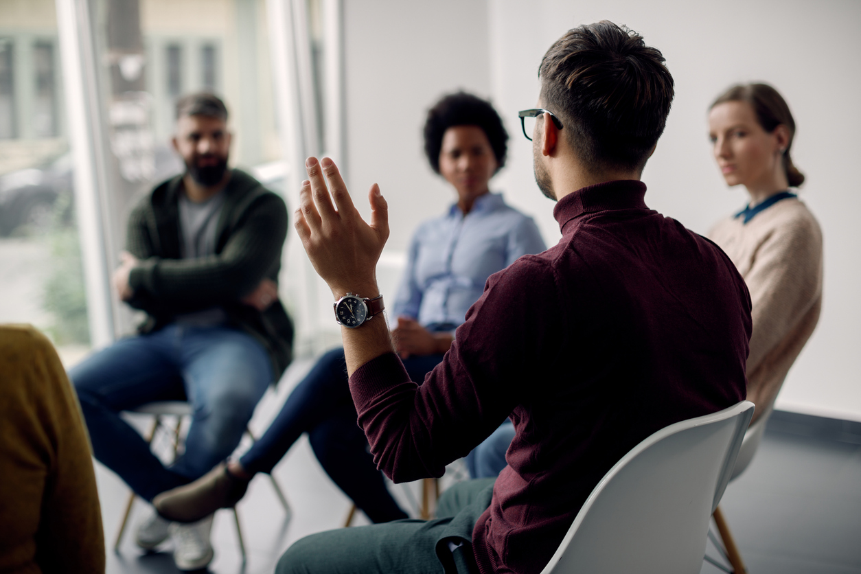 Rear view of man with raised hand on group therapy in medical center. Rear view of man raising his hand to ask a question while attending group therapy.
