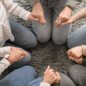 From an overhead view, a group of people sit in a circle on a grey rug, holding hands.