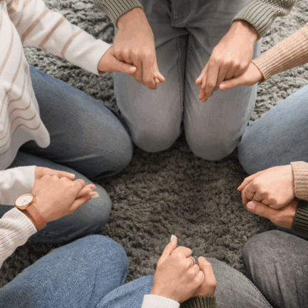 From an overhead view, a group of people sit in a circle on a grey rug, holding hands.