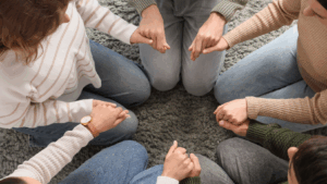 From an overhead view, a group of people sit in a circle on a grey rug, holding hands.