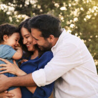 A joyful family moment with a father, mother in a blue dress, and their one-year-old son in blue clothes, hugging and smiling in a grassy natural setting with a tree in the background